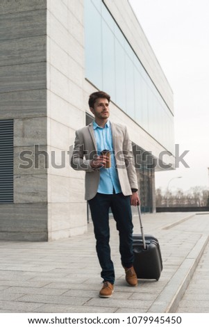 Only forward! Full length picture of the young handsome entrepreneur dragging a small luggage suitcase out the airport building and drinking coffee after arrival.