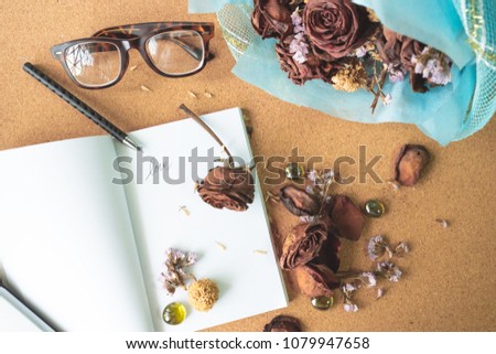 top view flat layer witerd old rusty brown roses and love word  on blank white book with blurry glasses and blue bouquet flower in background on crok wooden table, love forever, memory concept