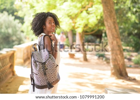Serious attractive young black woman holding rucksack in park, turning round, looking at camera and walking with blurred trees in background. Life balance and tourism concept. Back view.