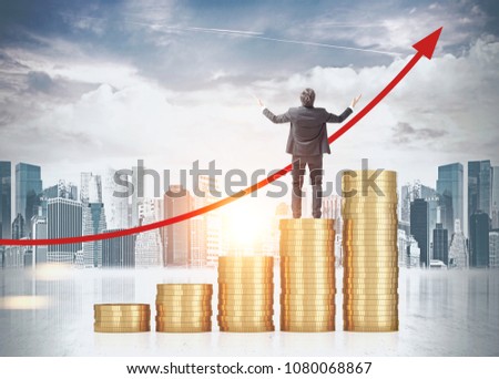 Rear view of a successful businessman celebrating a business victory. He is standing on a stack of huge coins looking at a growing graph in city sky. Toned image double exposure