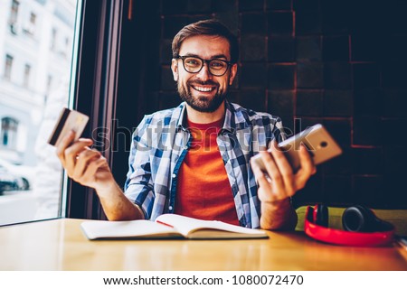 Half length portrait of happy bearded young man dressed in casual outfit smiling at camera while holding credit card and making money transaction via 4G internet connection on smartphone device
