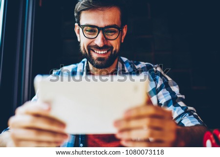 Positive bearded young man in eyeglasses for vision correction watching live stream on website on modern tablet via 4G internet.Cheerful male blogger enjoying transmission online on touch pad device