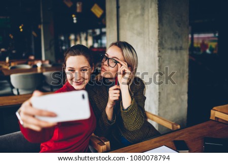 Two successful female bloggers in casual apparel with funny faces posing and making selfie photos for publication in social networks on modern smartphone during friendly meeting in cafe interior