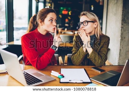 Brunette hipster blogger told secret to wondered colleague and showing sign shh during collaborating at laptop devices in coworking.Excited two best friends gossiping during studying break at netbooks Royalty-Free Stock Photo #1080078818