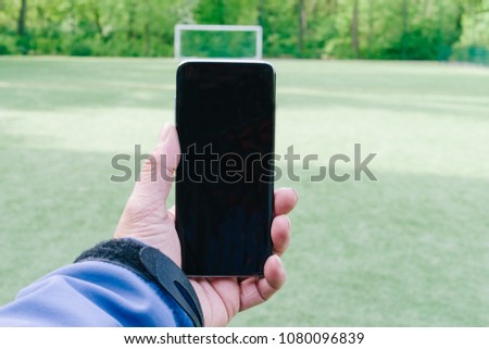 Man holding a smart phone in his hand with blank screen as copy space on grass soccer field in background.