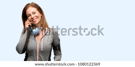 Middle age gym fit woman with workout headphones happy talking using a smartphone mobile phone isolated blue background