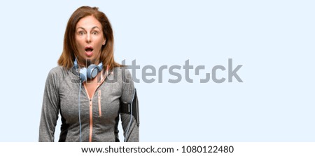 Middle age gym fit woman with workout headphones scared in shock, expressing panic and fear isolated blue background
