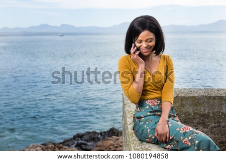 Beautiful black lady on her phone having a laugh while siting on a wall.