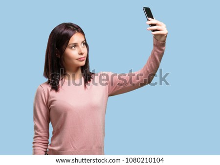 Young pretty woman confident and cheerful, taking a selfie, looking at the mobile with a funny and carefree gesture, surfing the social networks and internet
