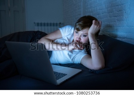 young beautiful, caucasian, red haired, internet addicted woman working and surfing bored, sleepless and tired on her laptop in bed in her bedroom late at night in dark room.