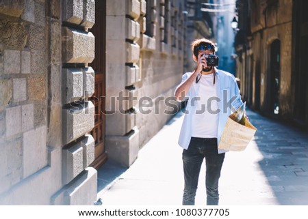 Young man tourist focusing and making photos on camera while strolling in narrow streets of old architectural city holding travel map in hand.Hipster guy taking pictures walking in urban setting