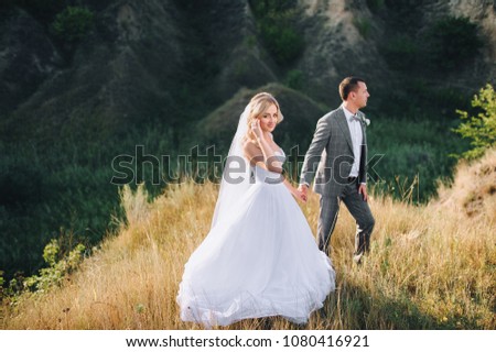 Beautiful newlyweds are standing, holding hands, in nature with the sun, against the background and hills. Evening wedding of the newlyweds with the control of the sunlight.