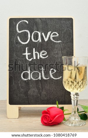 A blackboard with the words Save the date displayed with a glass of champagne and a red rose