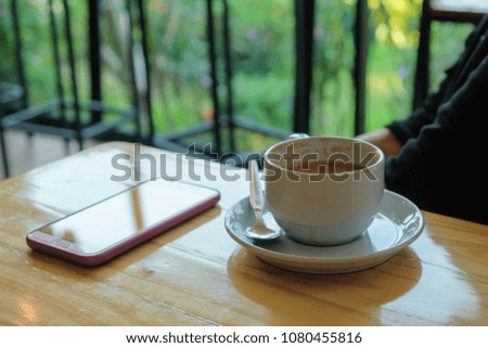 A white cup of coffee and a smartphone on wooden table beside green garden.