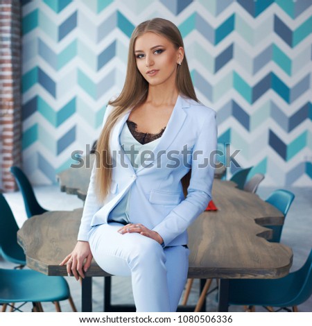 Young beautiful woman with long straight blond hair in blue suit sitting at wooden table in stylish modern interior. Natural lightning