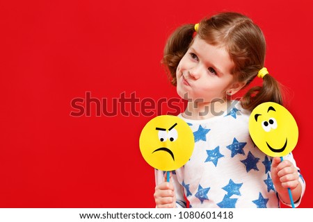 concept of children's emotions. child girl chooses between a sad and joyful smile on  colored red background
