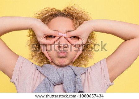 A picture of funny girl putting her fingers in a circle position and holding some air in her mouth. She looks funny but serious. Isolated on yellow background