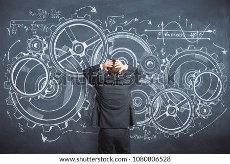 Back view of young pensive businessman standing on chalkboard background with cogwheel sketch. Risk and engineering concept 