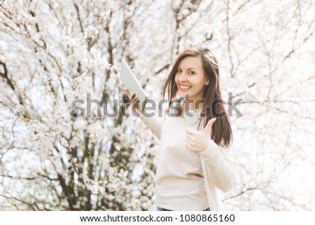 Young smiling beautiful woman in light casual clothes with headphones holding tablet pc computer showing thumb up in city garden or park on blooming tree background. Spring flowers. Lifestyle concept