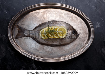 a large fresh sea fish. Fresh fish with lemon wedges and a rosemary branch on a silver platter on a dark background. Beautiful picture of food in a low key.