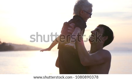 The father raises his son up in his arms, standing by the sea at sunset, the little boy spreads his hands to the sides. hd