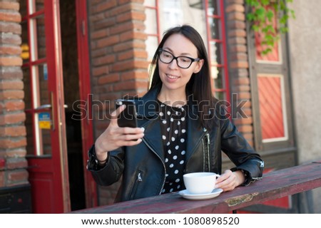 Beautiful brunette woman sitting in cafe, drinking tea and make a selfie on her phone. She is wearing black leather jacket and long dress .