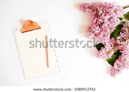 Cropped image of blank paper sheet on a clipboard, pink pen & lilac flowers in minimal arrangement on white table top background. Feminine flat lay composition w/ purple bouquet. Close up, copy space.