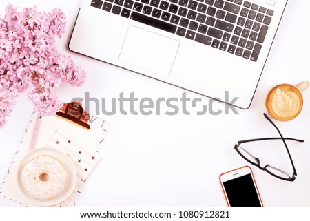 Feminine desktop, close up of laptop keyboard, blank clipboard, coffee & donut, lilac flowers. Notebook computer, blank screen cellphone, cappuccino cup, glasses eye wear, white background. Copy space
