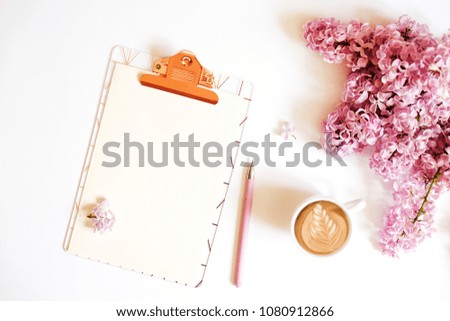 Blank paper sheet on a clipboard, cup of coffee, pink pen & lilac flowers in minimal arrangement on white table top background. Feminine flat lay composition w/ purple bouquet. Close up, copy space.