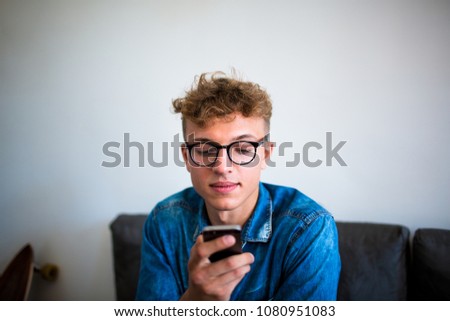 Stylish hipster guy using application on mobile phone, sitting in cafe. Fashionable man in glasses reading text message in social network via cellphone. Young male watching video on smartphone 