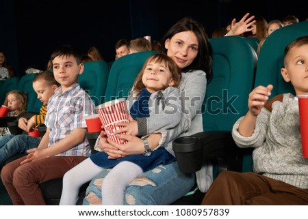 Spectators sitting in comfortable chairs in movie theatre, watching film. Beautiful woman and cute little girl posing at camera, holding paperbag with popcorn in hands. Children drinking fizzy drinks.