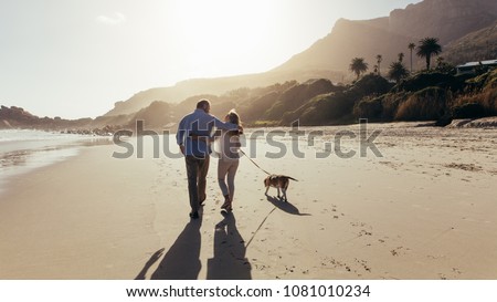 Mature couple strolling along the beach with their dog. Rear view shot of loving mature couple on the beach with dog. Royalty-Free Stock Photo #1081010234
