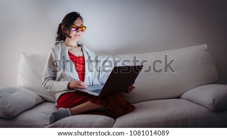Young woman with blue light blocking glasses (yellow lenses) working with laptop on white sofa Royalty-Free Stock Photo #1081014089