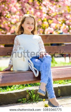 Girl on smiling face sits on bench, sakura tree on background, defocused. Cute child with bag or backpack enjoy sunny spring day. Girl relaxing during walk in park near cherry blossom. Relax concept.