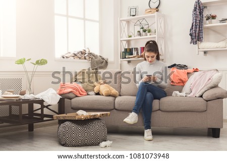 Desperate helpless woman sitting on sofa in messy living room. and chatting on mobile, surrounded by many stack of clothes. Disorder and mess at home, copy space Royalty-Free Stock Photo #1081073948