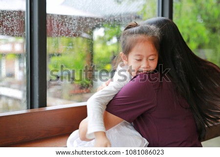 Beautiful Asian mother and her daughter embracing with love near a window while raining day.