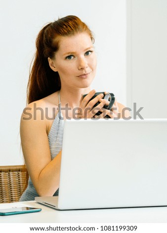 Photo of a beautiful young woman with red hair sitting with a laptop and drinking coffee.