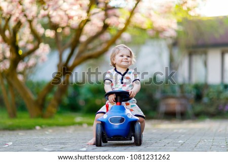 Cute little baby girl playing with blue small toy car in garden of home or nursery. Adorable beautiful toddler child with blossoming magnolia on background