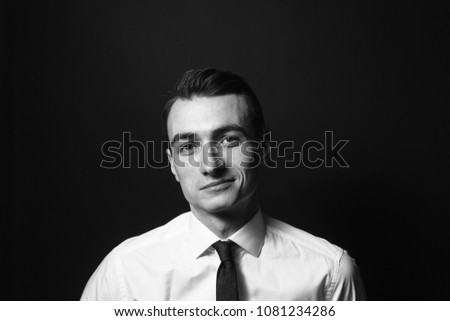 Close up black and white portrait of a young man in a white shirt and black tie, smiling while looking at the camera, against plain studio background