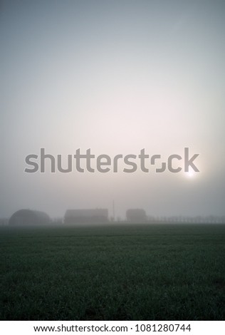 The Silhouettes of Countryside Buildings in an Early Morning  with a Field in a Foreground and a Heavy Mist Covering the Background. Moody Photo with a Text Space