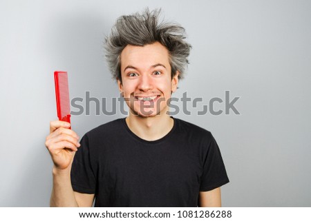 Unshorn and unshaven young guy holds comb on gray background.