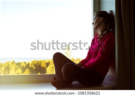 Portrait of a beautiful young woman sitting on a window sill with legs crossed, and enjoying the view from the window.