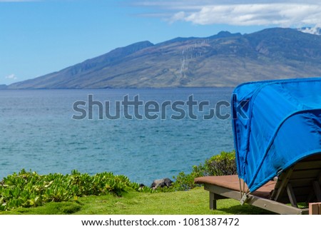 Blue covered cabana sits on green grass on the edge of the ocean, looking out over blue waters, of Maui Hawaii, to the distant mountain with a row of windmills 