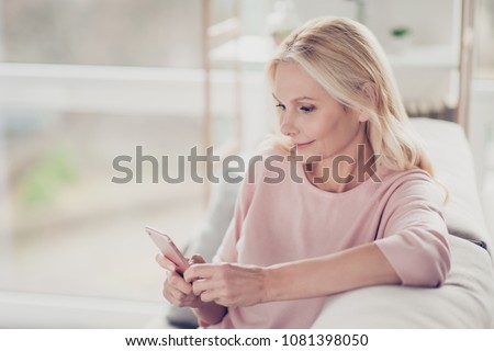Charming, pretty, stylish, modern grandmother sitting on sofa in living room at home, doing online shopping through 3G internet, having smart phone in hands