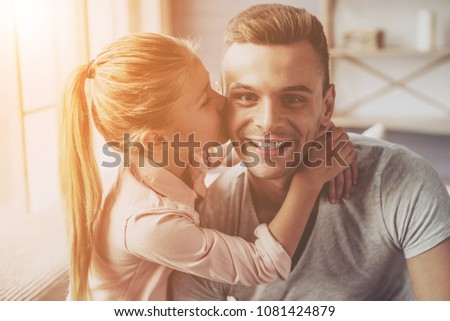 Portrait of handsome young father and his little daughter hugging and smiling while sitting on sofa at home. Girl is kissing her dad in cheek