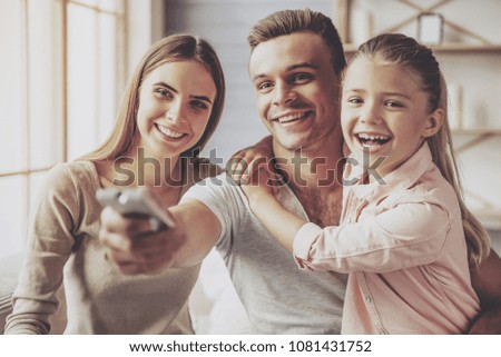 Beautiful happy young parents and their little daughter watching TV and smiling while sitting on sofa at home