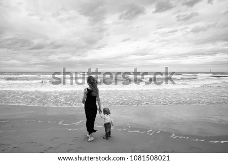 Mother in black dress and son in shirt jeans back view standing on sea shore beach wet sand watching waves running on coast on windy murky day on seascape background, horizontal picture