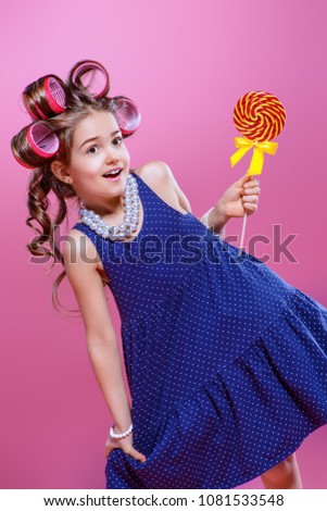 Portrait of a pin-up little girl with curlers in her hair holding lollipop. Studio shot over pink background. Kid's fashion. 