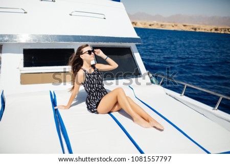 Beautiful young woman in bikini adjusting eyewear and smiling at camera while lying on the deck of yacht