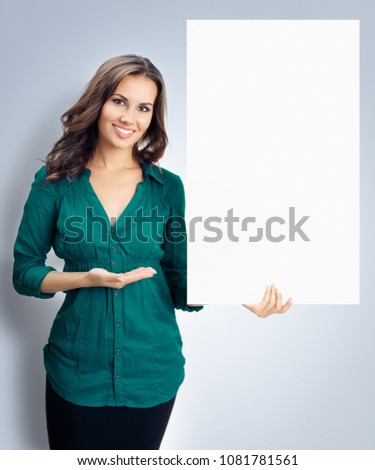 Happy smiling young woman in green confident clothing showing blank signboard with copyspace empty area for some text or slogan, over grey background. Success in business and advertising concept.